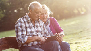 Senior couple sitting on a park bench on sunny autumn day with tablet and relaxing.