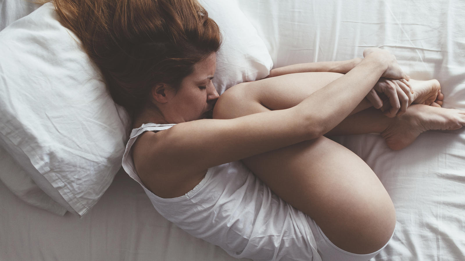 Eine junge Frau liegt im Bett und hält sich vor Schmerzen ihre Beine fest.