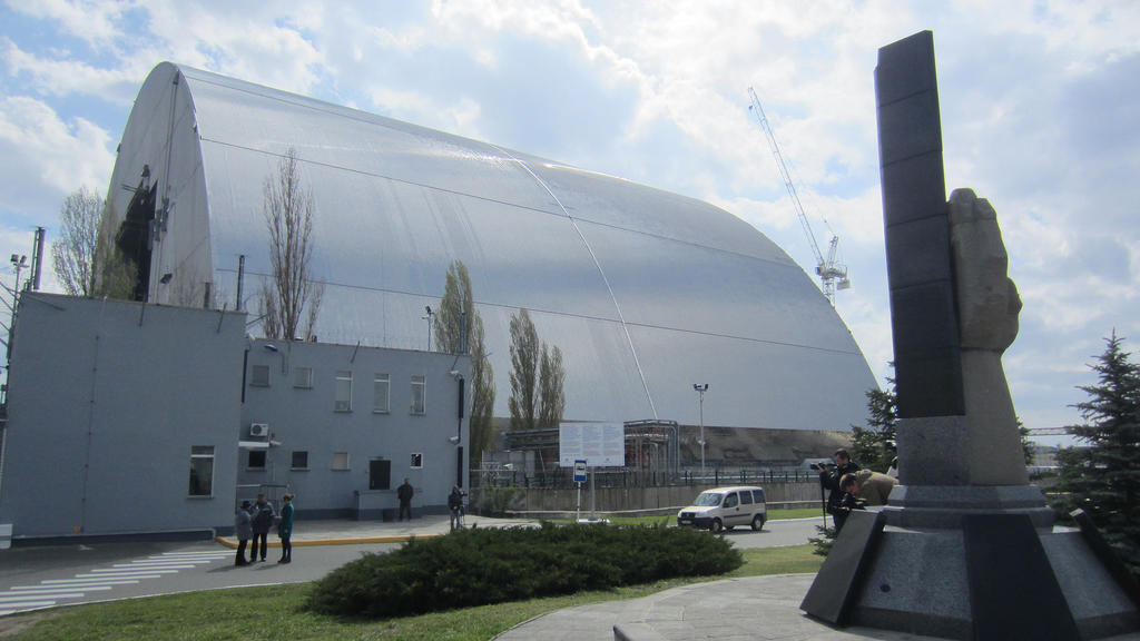 ARCHIV - Ein Mahnmal für die Opfer der Reaktorkatastrophe ist am 22.04.2016 vor der neuen Hülle des Unfallreaktors in Tschernobyl, Ukraine, zu sehen. Der neue Schutz ist über 100 Meter hoch und wiegt mehr als 30.000 Tonnen.   Foto: Andreas Stein/dpa 