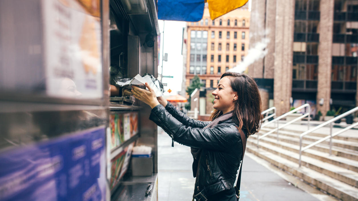 A beautiful Caucasian adult woman buys some lunch from a New York city food cart, a smile on her face.  She wears modern stylish clothing with darker and black colors.  Horizontal image with copy  space.