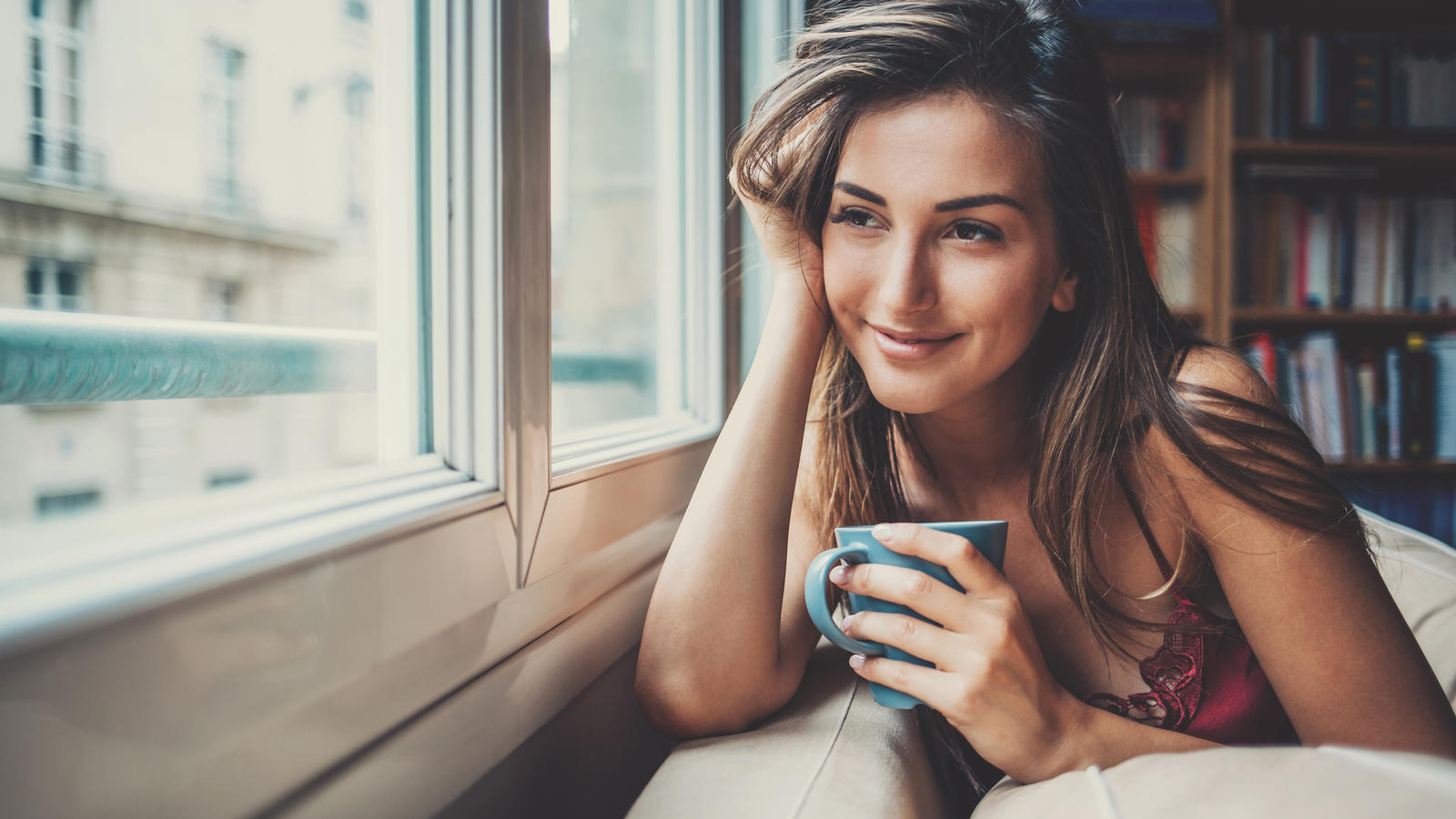 Junge Frau sitzt mit Kaffeetasse am Fenster und schaut hinaus