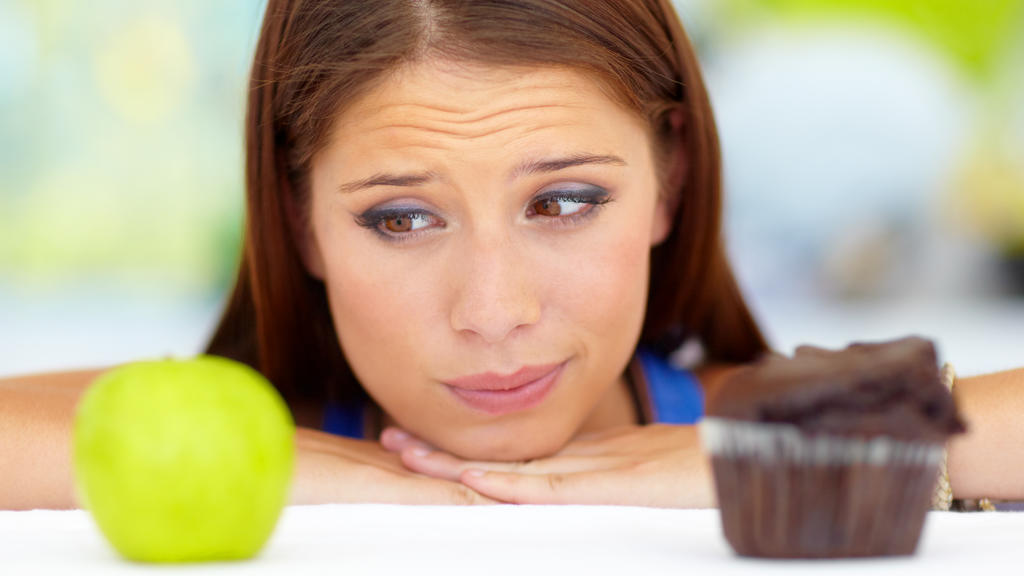 Eine junge Frau kann sich nicht zwischen einem Apfel und einem Muffin entscheiden. Gesund vs. ungesund.
