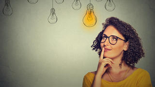 Portrait thinking woman in glasses looking up with light idea bulb above head isolated on gray wall background