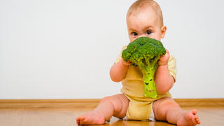 Cute baby eating broccoli sitting on floor indoors