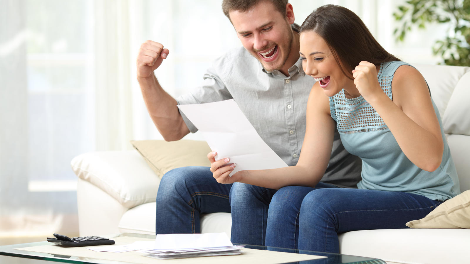 Excited couple reading a letter together sitting on a sofa in the living room at home