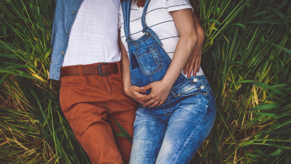 Photo of a young couple, soon to become parents, resting in the middle of the field on a beautiful spring day while expecting a new life to begin. Baby bump is in a focus, while parents are out of the frame