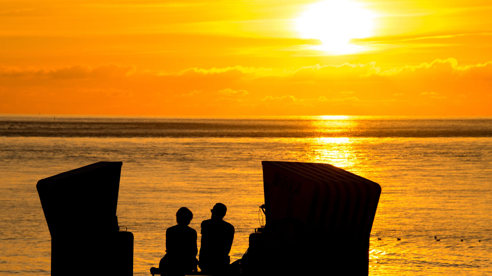 ARCHIV - ILLUSTRATION - Zwei Menschen sitzen am 26.07.2016 in Norderney (Niedersachsen) im Sonnenuntergang zwischen zwei Strandkörben. «Hygge» steht im Dänischen für die kleinen Freuden im Alltag, die die Lebensqualität erhöhen. (zu dpa «Der Hype um 