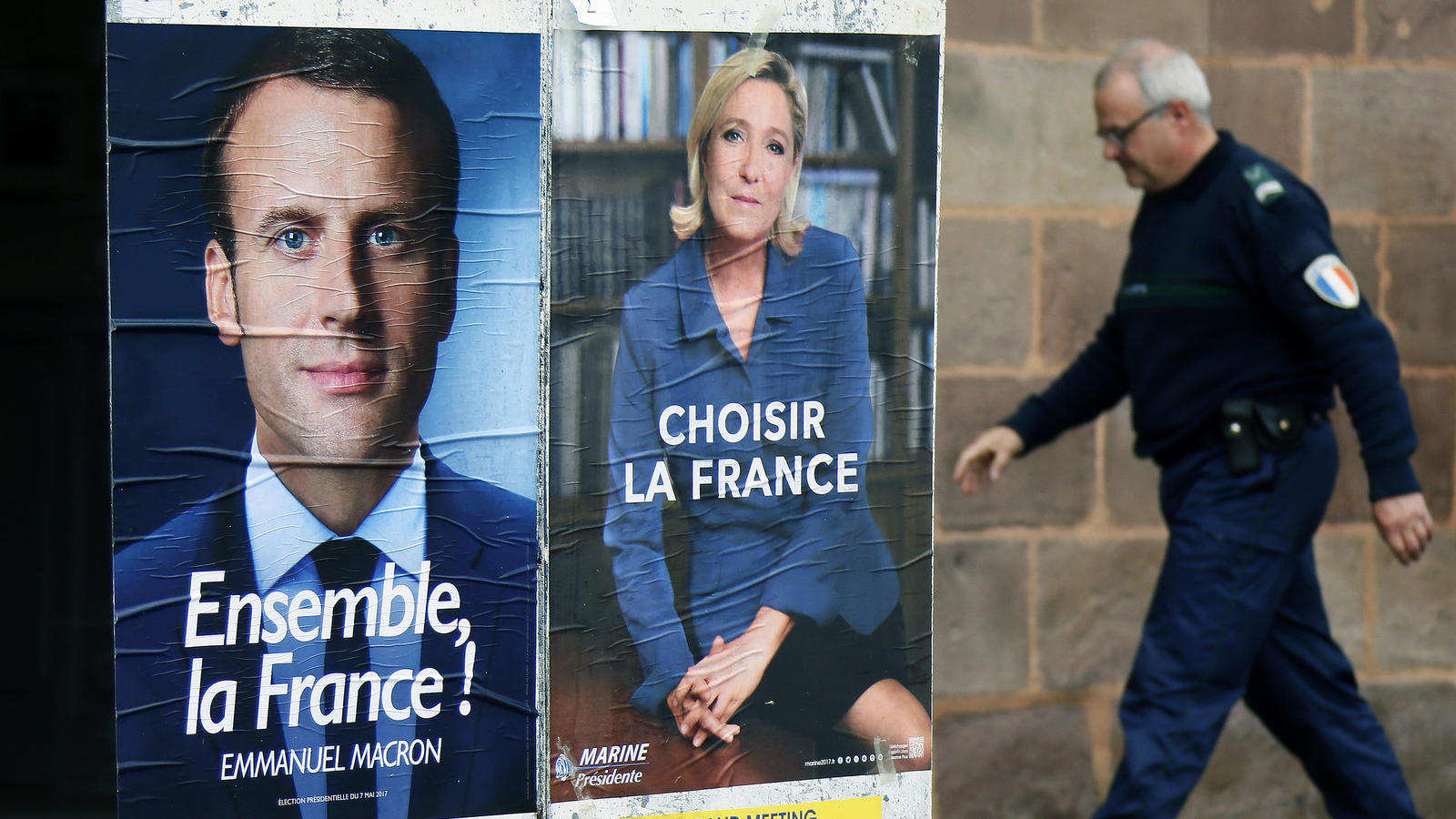 Ein Polizist geht am 05.05.2017 in Saint Jean Pied De Port (Frankreich) an den Wahlplakaten von Emmanuel Macron (l) und Marine Le Pen (r) vorüber. Foto: Bob Edme/AP/dpa +++(c) dpa - Bildfunk+++