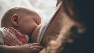 A newborn baby sleeps in her mother's arms while breastfeeding from her mother. The baby's face in in focus as the mother is unrecognisable.