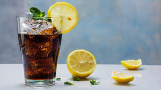 glass of cola or ice tea with ice cubes, lemon slice and peppermint garnish, against a blue wall