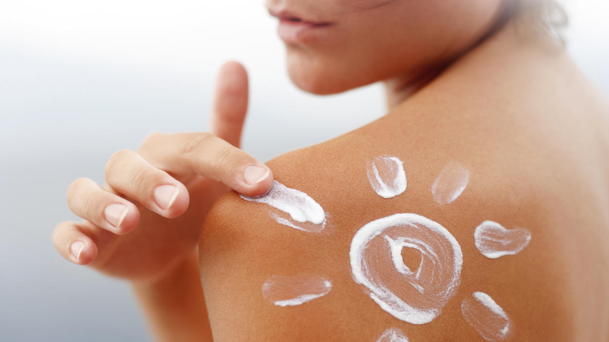 Frau cremt sich die Schulter mit Sonnencreme ein. Die Creme stellt eine Sonne dar.