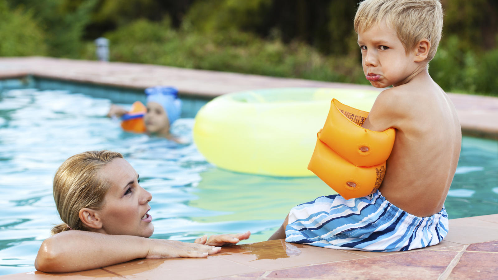 Little boy wearing water wings sitting by the swimming pool looking angry and sad, while mother talks to him.