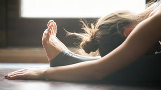 Young woman practicing yoga, sitting in Seated forward bend exercise, paschimottanasana pose, working out, wearing sportswear, grey pants, bra, indoor, home interior background, close up