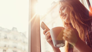Smiling woman using smartphone and drinking coffee in the morning