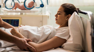 Young woman lying in hospital bed with her eyes closed after childbirth while unrecognizable person is consoling her.