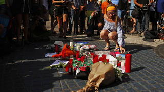 Eine Frau stellt am 18.08.2017 auf dem Gehweg nahe dem Bereich auf Flaniermeile Las Ramblas in Barcelona (Spanien), auf dem Tags zuvor ein Lieferwagen in eine Menschenmenge gefahren war, eine Postkarte zu Blumen und Kerzen. Bei dem Terroranschlag wurden mehrere Menschen getötet und viele verletzt. Die IS-Terrormiliz hat den Terroranschlag für sich reklamiert. Foto: Francisco Seco/AP/dpa +++(c) dpa - Bildfunk+++