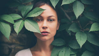 Portrait of beautiful fashion model looking at camera from the green leaves of big plant. She has no make up. She wears a white shirt and looks amazing.