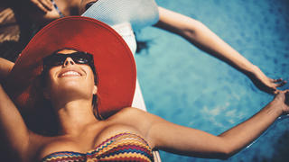 Closeup of two mid 20's women sunbathing by a pool side on hot summer day. Both wearing hats and sunglasses and holding hands above water. Closeup, top view.