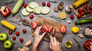 Female hand cut tomatoes on rustic kitchen table, around lie ingredients, vegetables, fruits, and spices, Healthy foods, cooking and vegetarian concept.