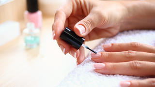 Closeup of adult caucasian unrecognizable woman painting her fingernails with nail polish product. Her hands are placed on a white towel. blurry cosmetic products in background.