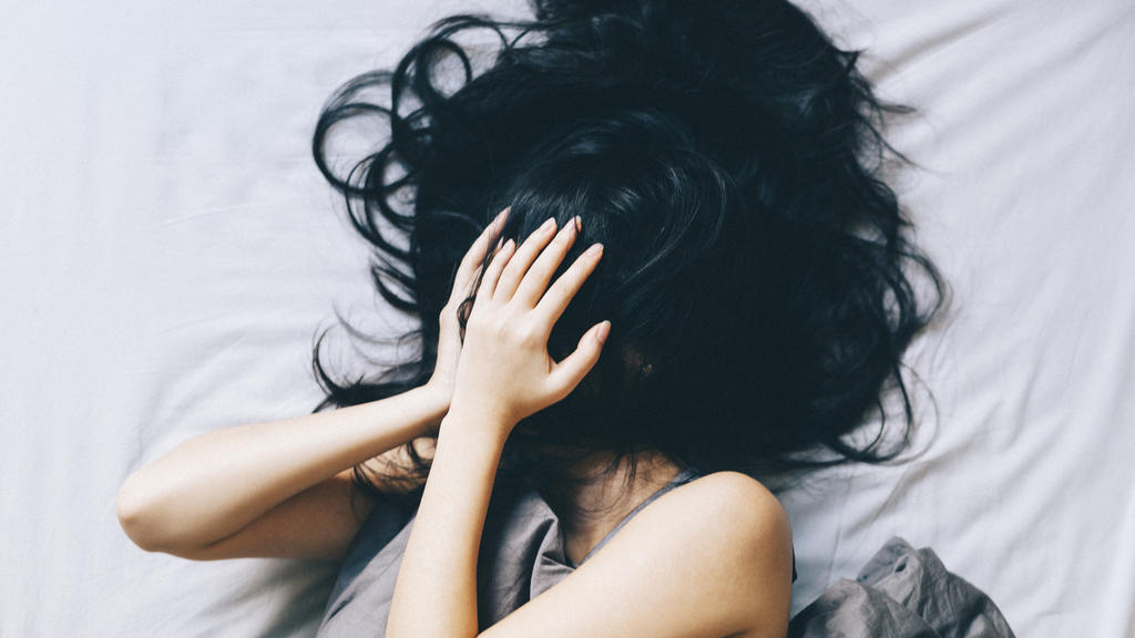Eine junge Frau liegt im Bett, scheint aber durch die Hände vor ihrem Gesicht einen sehr gestressten Eindruck zu machen.