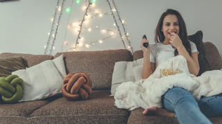 Good-looking girl is wearing blue jeans and white t-shirt. She feels relaxed. While she is watching TV shows she is eating popcorn.