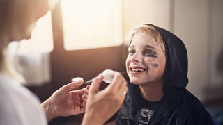 Little boy aged 8 preparing for halloween. Mother is painting the boy face and the boy is smiling at his mother.