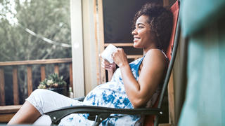 A beautiful African American woman in the 3rd trimester of her pregnancy rests in a recliner on her porch, enjoying a hot drink while anticipating her upcoming child birth.  Horizontal image with copy space.