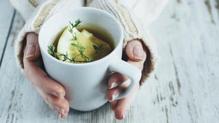 Human hands holding cup of tea with thyme herb and lemon slices on a wooden table,.