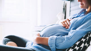Beautiful pregnant woman sitting at bed and holds hands on belly in bedroom at home. Pregnancy, parenthood, preparation and expectation concept. Close-up, indoors.