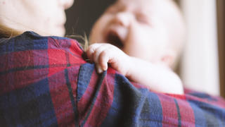 Newborn baby wearing red and white striped bodysuit, in her mother's arms, crying. Mother wearing red and blue plaid shirt and blue jeans, holding baby with care. Baby holding mothers shirt, focus on baby's hand.