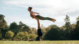 Fit young couple doing acro yoga in park. Man lying on grass and balancing woman in his feet.