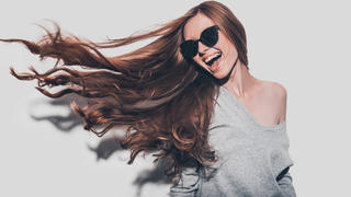 Attractive young smiling woman in sunglasses and with tousled hair looking away while standing against grey background