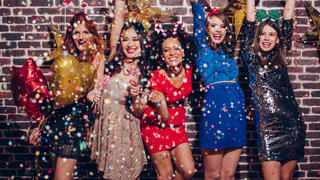 Group of beautiful young girls celebrating New Year. Standing in front of a brick wall, laughing and dancing. Holding lollipops. Wear elegant dresses. Confetti in the air. Room is decorated with star and heart  shaped balloons.