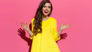 Happy beautiful young woman with curly long brown hair posing in yellow dress. Three quarter length studio shot on pink background.