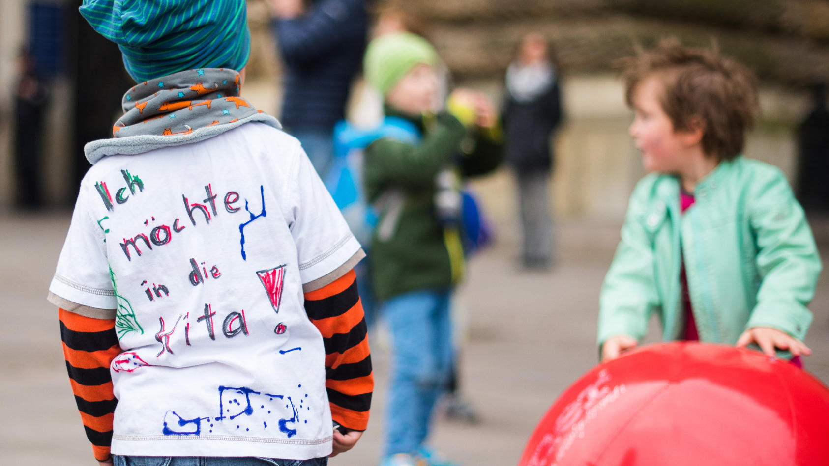 ARCHIV - Ein Kind mit einem T-Shirt mit der Aufschrift «Ich möchte in die Kita» steht am 28.05.2015 in Hamburg während einer Aktion von Eltern zum Ende des Kita-Streiks vor dem Rathaus neben Kindern, die mit Bällen spielen. Foto: Daniel Bockwoldt/dpa