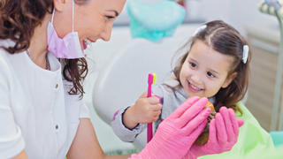Smiling girl sitting in dentist chair with her pediatric dentist showing her teeth model and teaching her how to brush teeth. Dentistry, oral hygiene concept